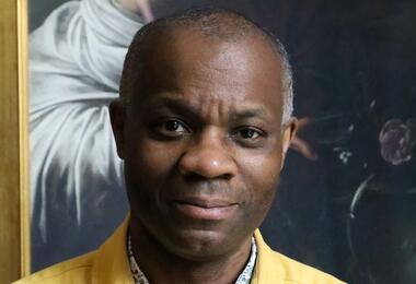 A man wearing a yellow shirt poses in front of a vibrant painting, showcasing his appreciation for art.