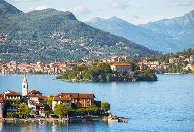 A serene view of Lake Maggiore, showcasing its calm waters surrounded by lush green hills and distant mountains.