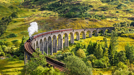 The Jacobite steam train crosses the Glenfinnan Viaduct, surrounded by lush green hills in Scotland.