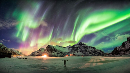 A man stands before a mountain, illuminated by a vibrant green aurora borealis in the night sky.
