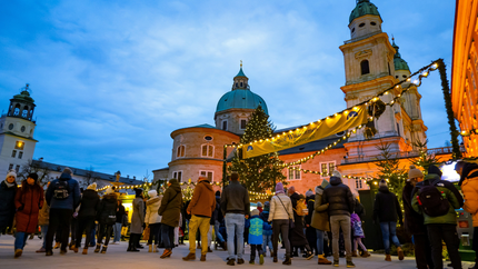 People stroll in front of a church, with a large Christmas tree adorned with lights and decorations nearby.