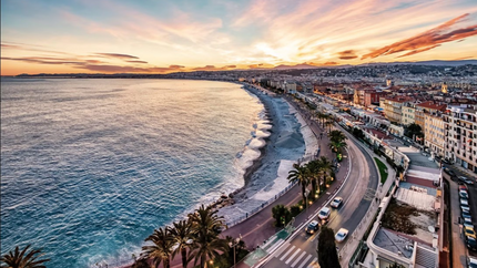 Sunset over the beach and ocean in Nice, France, with vibrant colors reflecting on the water and silhouettes of palm trees.