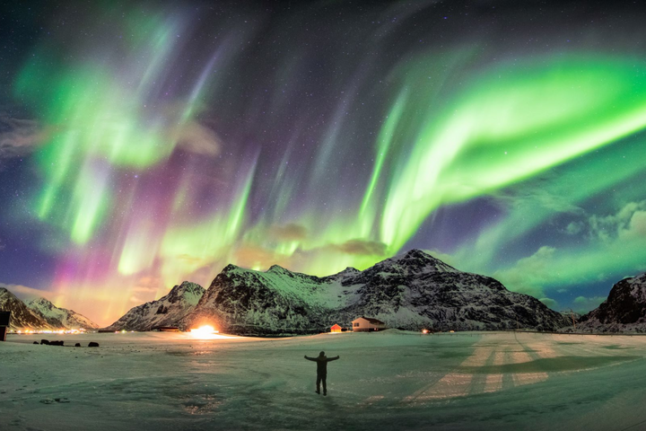 A man stands before a mountain, illuminated by a vibrant green aurora borealis in the night sky.