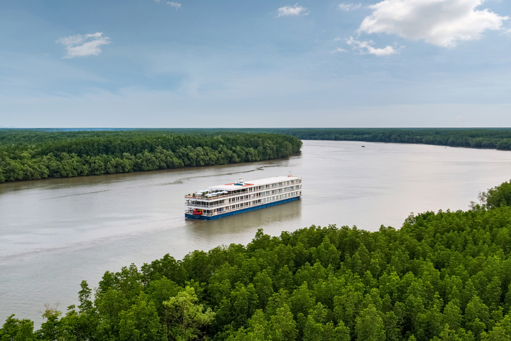 A cruise ship glides down a river, flanked by lush green trees on both sides.