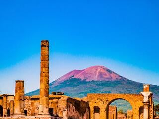 Ruins of Pompeii with ancient structures in the foreground and Mount Vesuvius towering in the background.