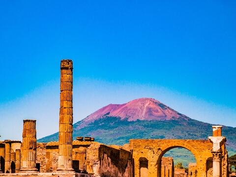Ruins of Pompeii with ancient structures in the foreground and Mount Vesuvius towering in the background.