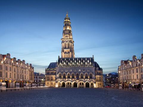 A bustling city square in Arras, featuring the iconic cathedral surrounded by historic buildings and visitors.