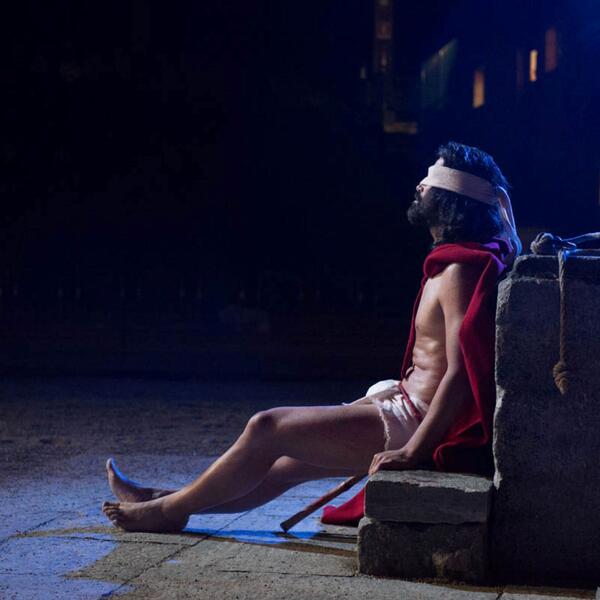 A character from the Sordevolo Passion Play, dressed in a red robe, sits on a stone, reflecting the play's dramatic themes.